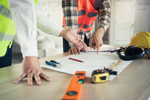 This image shows a general contractor and a business owner wearing high visibility vests discussing floorplans that are on the table in front of them. On the table there are a variety of tools and equipment including a tape measure and pliers and ear defenders and a hard hat.