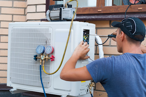 This image shows a tradesperson using a screwdriver and other equipment to fix or install a HVAC system on the exterior of a residential house's wall.
