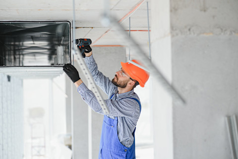 This image shows a HVAC engineer installing a heat recovery ventilation system in a commercial building. He is wearing a hard hat, gloves and safety glasses and is using an electric screwdriver or drill.
