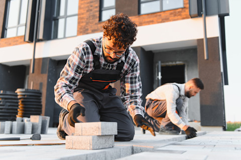 Two construction workers kneel while laying outdoor paving. They work together to align the stones.