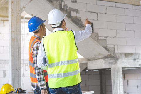 This image shows a general contractor and an engineer in a building that is being renovated. They are wearing high visibility vests, one in orange and one in yellow, as well as hard hats. They are pointing at something and discussing it.