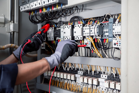 An electrician performs electrical testing and maintenance in an industrial setting. Cables and testing equipment surround the workspace.