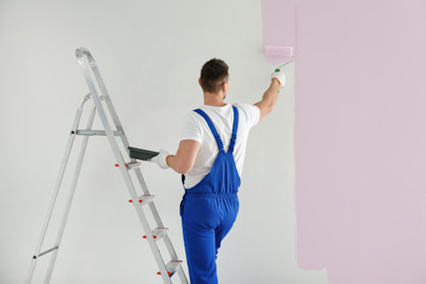 This image shows a painter and decorator dressed in overalls and using a roller brush to paint a wall pale pink.