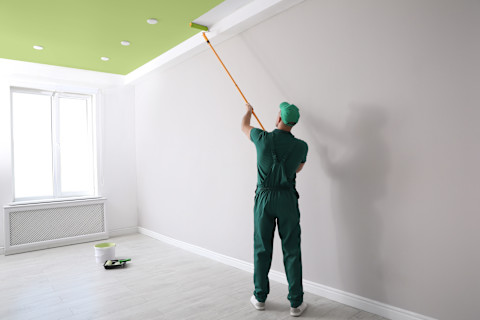 A man paints a ceiling green inside a room. The ceiling surface is partially covered.