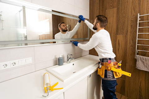 A worker mounts a mirror on a bathroom wall. The mirror is positioned above a sink in a clean, modern interior.