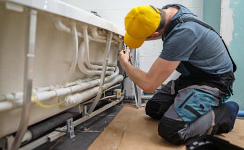 A technician services a pipe system beneath a bathtub with a hydromassage unit.