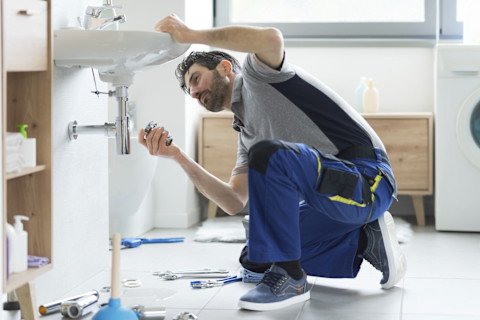 A plumber sitting under a sink with work clothes on to repair a sink